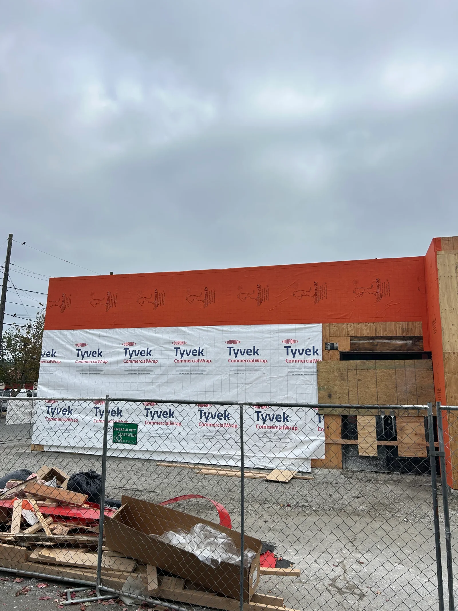 Commercial building with red exterior, Tyvek wrap, plywood sheathing, and protective fencing during renovation